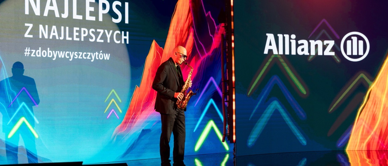 Paweł Brzeźnicki gra na saksofonie na scenie gali firmowej Allianz, przed ścianą LED z logo