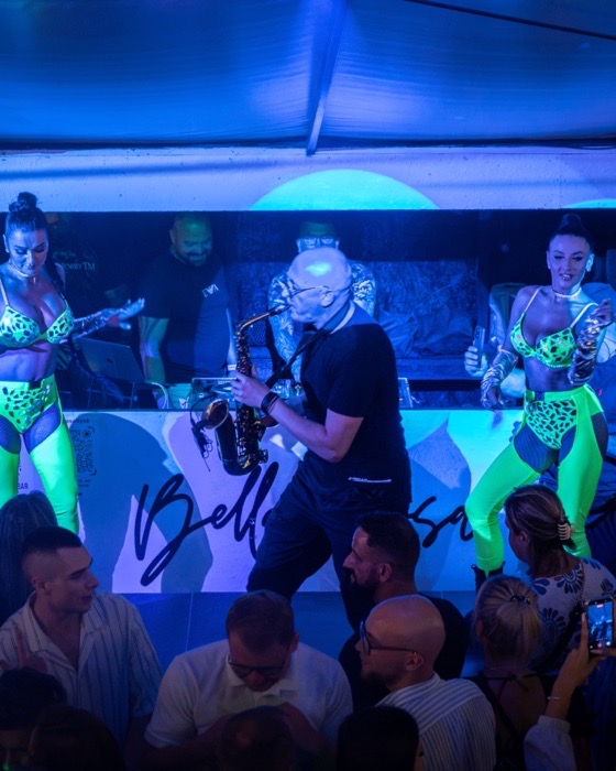 Saksofonista gra na scenie plenerowego klubu, w tle DJ przy konsolecie i tancerki