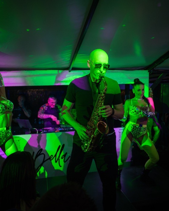 Saksofonista w zielonym świetle scenicznym, w tle DJ przy konsolecie i tancerki